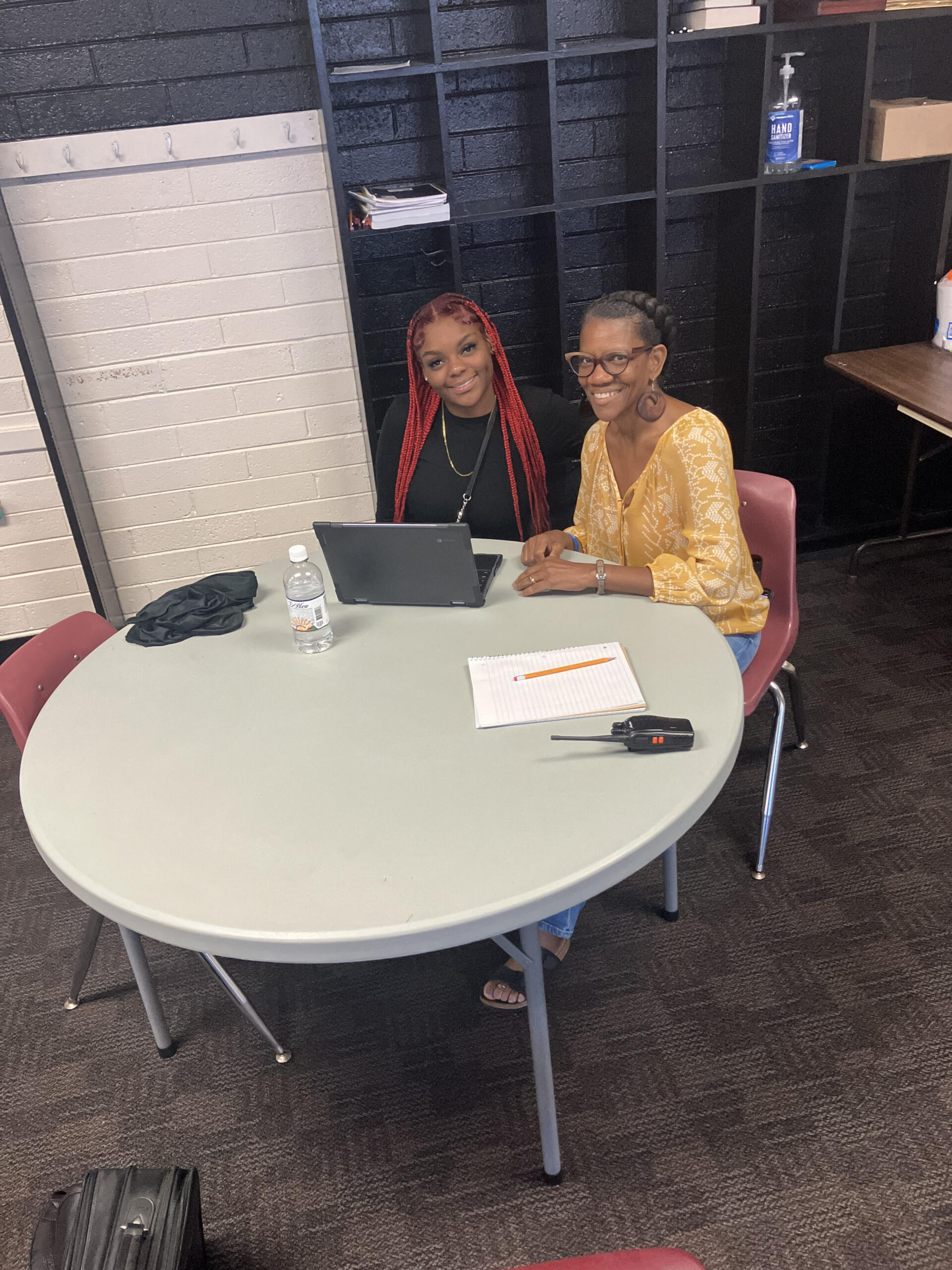 Two women sit at a round table with a laptop, notebook, and water bottle, smiling at the camera.